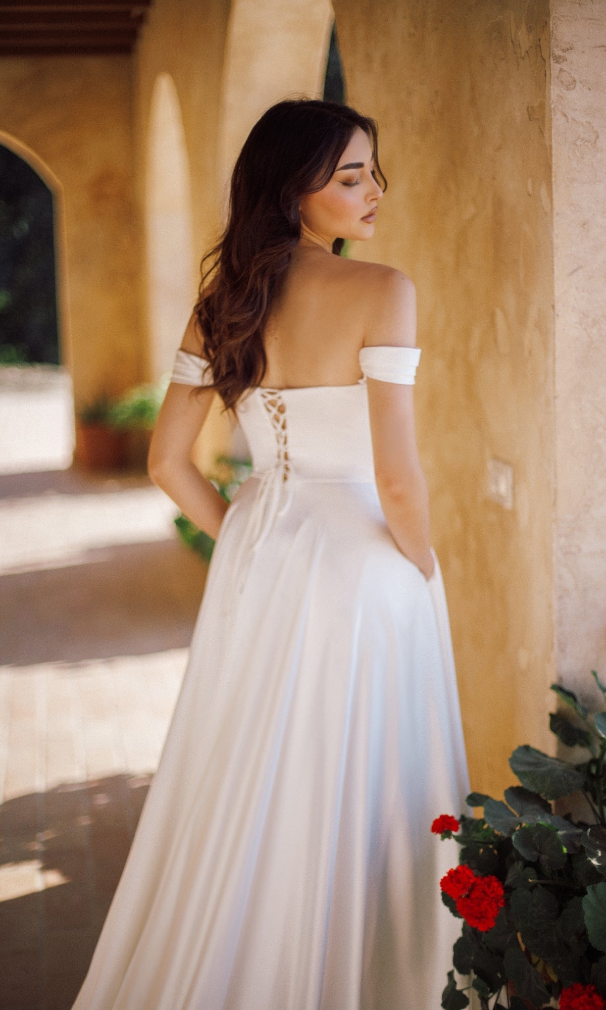 A woman wearing the Tina Holly TE565 Long Formal Dress with an off-the-shoulder neckline and lace-up back stands on a sunlit stone veranda near red flowers, facing away from the camera.